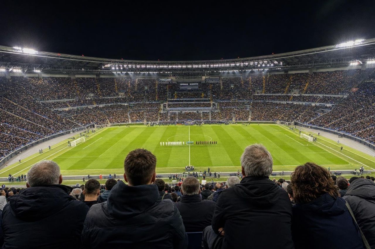 Pełny stadion nocą widziany z górnej trybuny, drużyny ustawione na boisku (obraz wygenerowany przez AI)